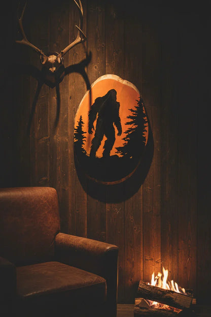 Bigfoot sunset wood slice art displayed on dark wood paneled man cave wall with antlers and amber firelight