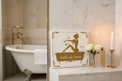 Salle de bain white wood sign with gold silhouette leaning on a marble bathroom shelf with white roses, bath oil, and a lit candle